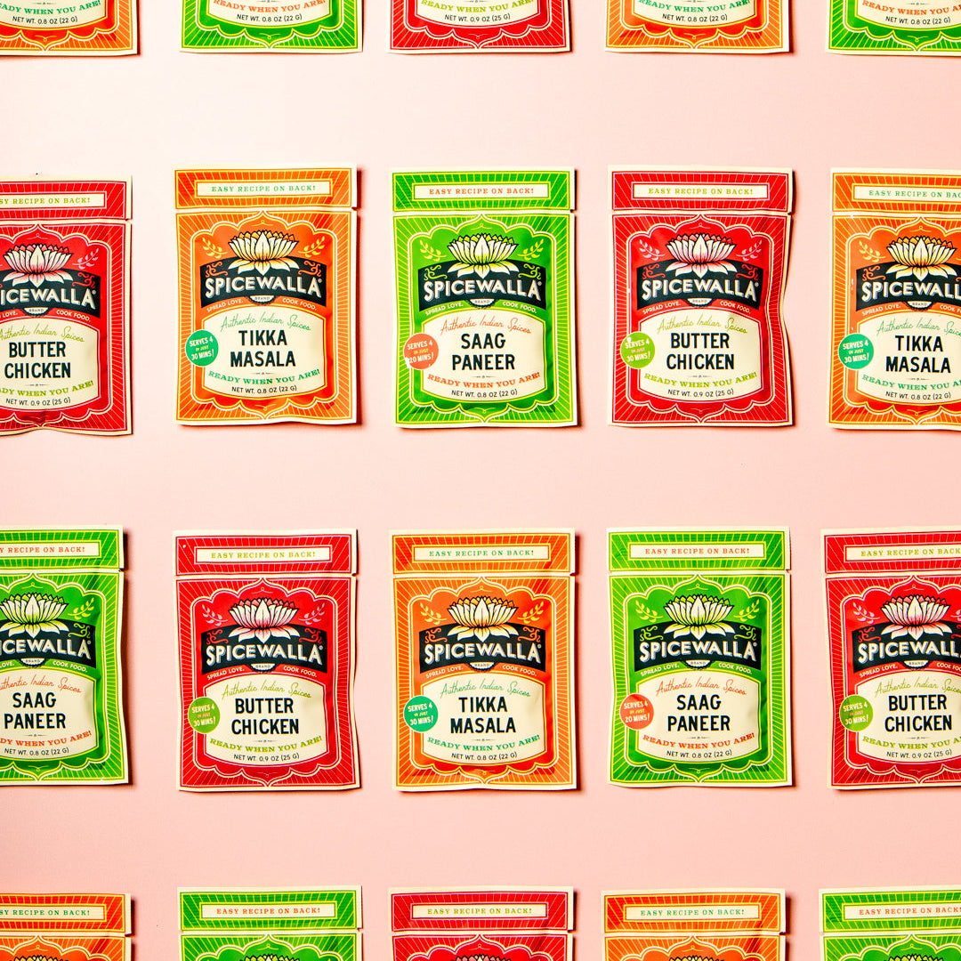 A grid of Spicewalla brand seasoning packets arranged in four rows on a pale pink background. The packets include Butter Chicken, Tikka Masala, and Saag Paneer flavors in red, orange, and green packaging.