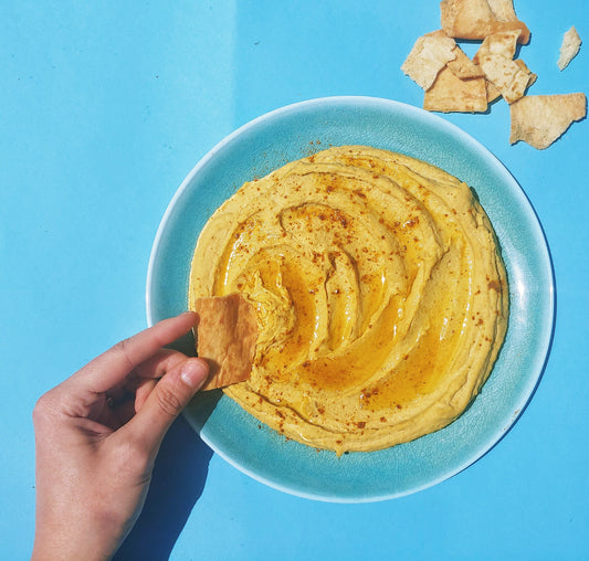 blue background with a hand dipping a pita chip into a bowl of creamy hummus