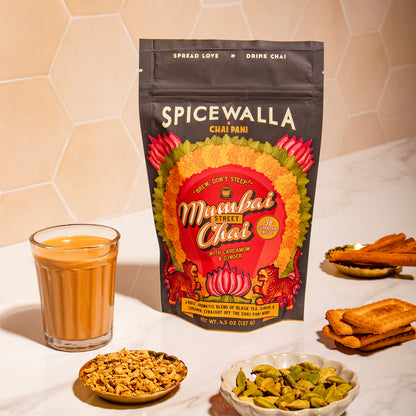 Spicewalla chai package with a glass of chai, snacks, and a bowl on a white surface.