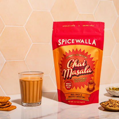 Spicewalla Chai Masala package with a glass of chai and cookies on a beige hexagonal tile background