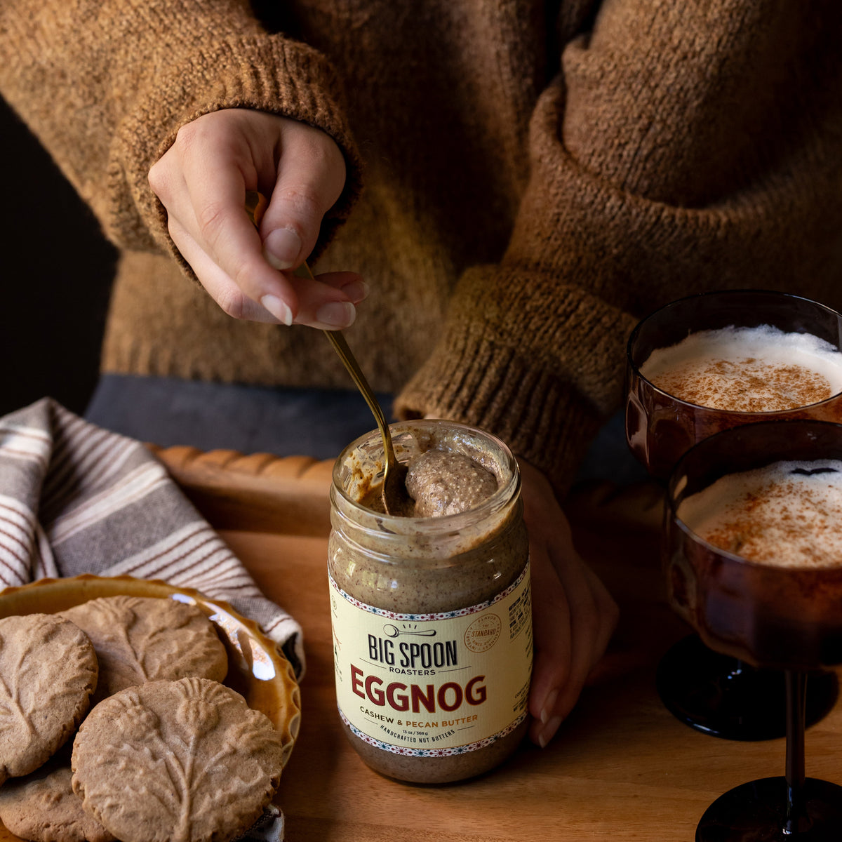 A person in a brown knit sweater spoons egg nog from a glass jar labeled Big Spoon Egg Nog onto a wooden tray, with cookies on the left and two glasses of egg nog on the right.