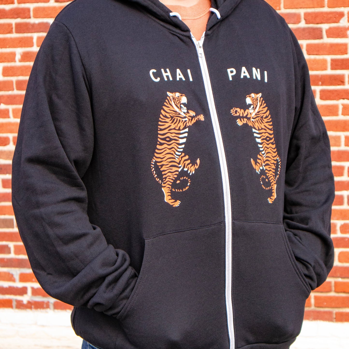 Person wearing a black hoodie with tiger graphics and 'Chai Pani' text against a brick wall.