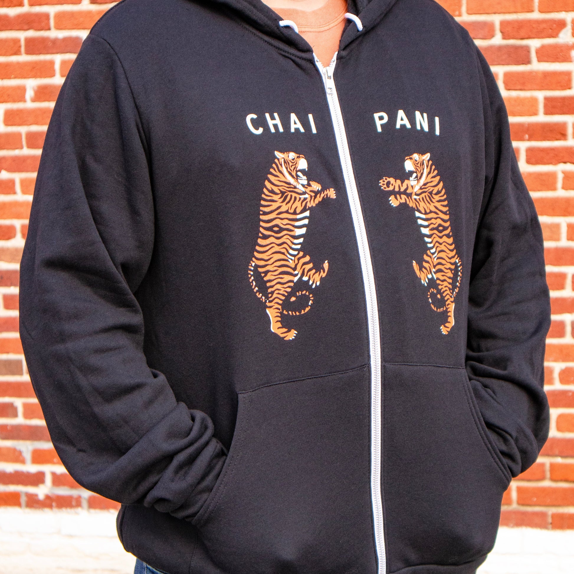 Person wearing a black hoodie with tiger graphics and 'Chai Pani' text against a brick wall.