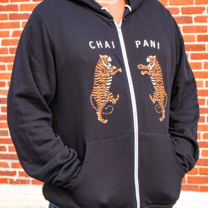 Person wearing a black hoodie with tiger graphics and 'Chai Pani' text against a brick wall.