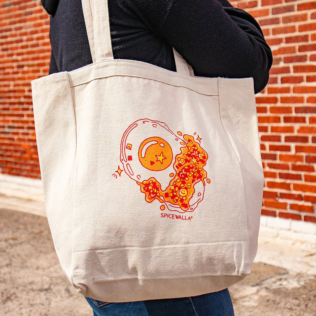 Person holding a beige tote bag with a colorful design and 'SPICE WALLA' branding against a brick wall.