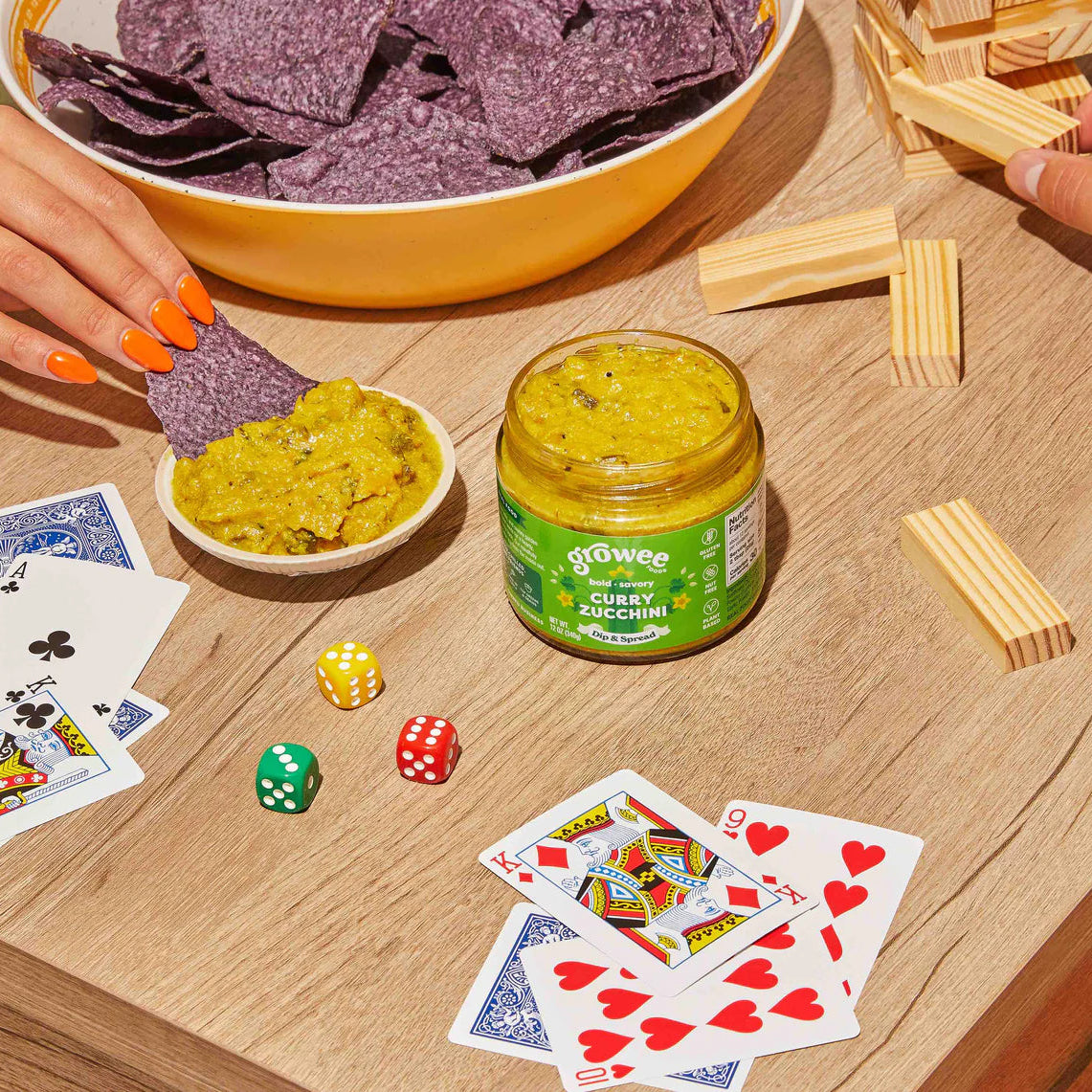 Game night setup with chips, guacamole, dice, cards, and Growe's Curry Dip on a wooden table.