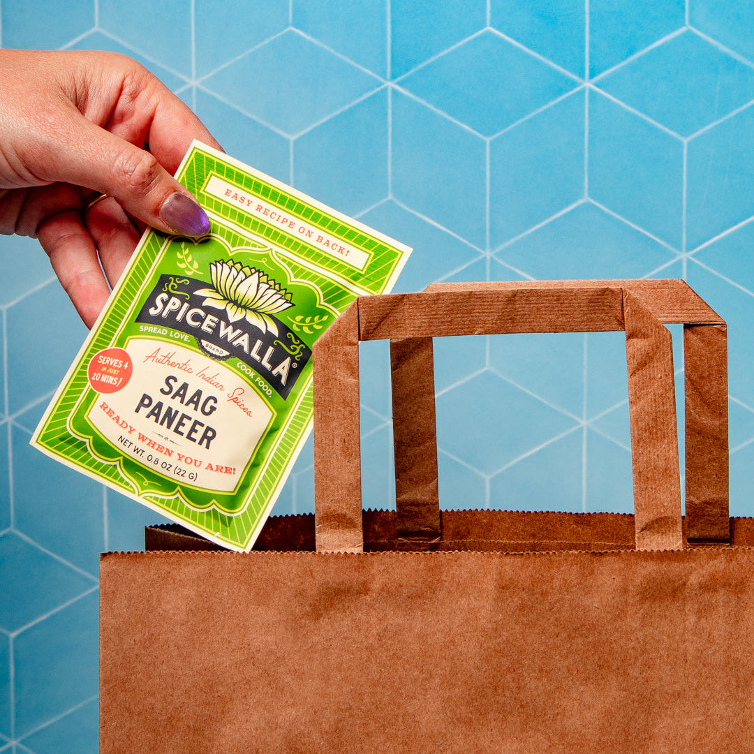 A hand with purple nail polish holds a green and white packet of Spicewalla Saag Paneer above a brown paper shopping bag against a blue geometric background.