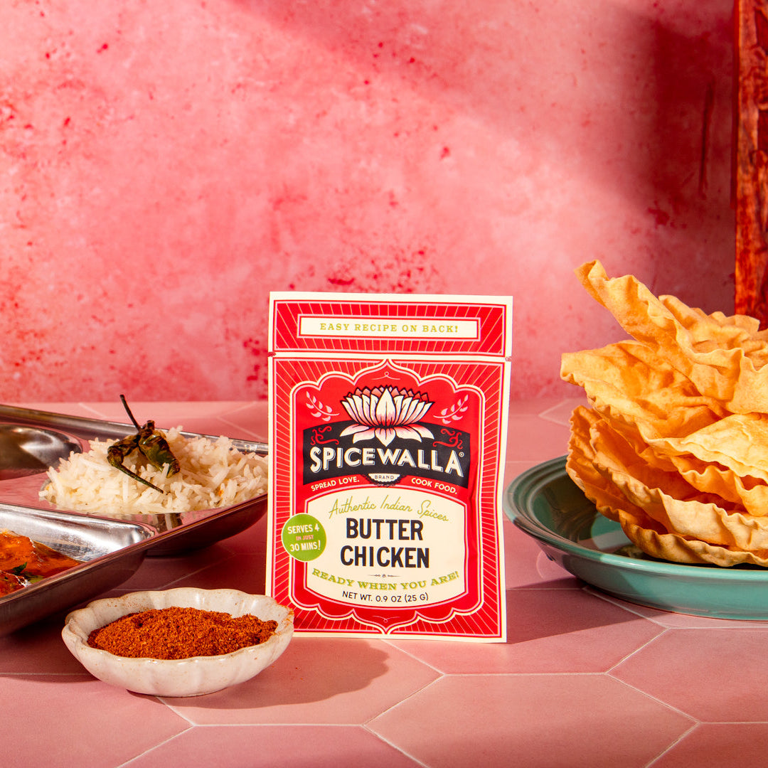 A packet of Spicewalla Butter Chicken seasoning mix stands on a pink surface, surrounded by prepared food including a tray of chicken, rice, a small bowl of red spice, and a plate of golden crispy crackers against a textured pink background.