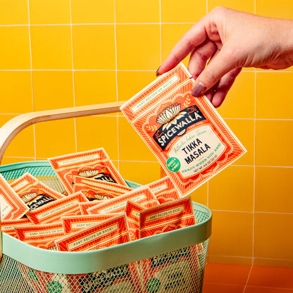 A hand holds a packet of Spicwalla Thika Masala spice mix above a mint green mesh basket filled with multiple packets, set against a bright yellow tiled background.