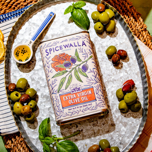 A tin of Spicewalla Extra Virgin Olive Oil is displayed on a silver tray, surrounded by fresh olives, basil leaves, sliced oranges, and a small dish of oil with seeds, set on a woven mat over a green tiled surface.