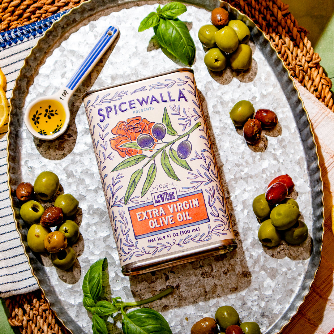 A tin of Spicewalla Extra Virgin Olive Oil is displayed on a silver tray, surrounded by fresh olives, basil leaves, sliced oranges, and a small dish of oil with seeds, set on a woven mat over a green tiled surface.