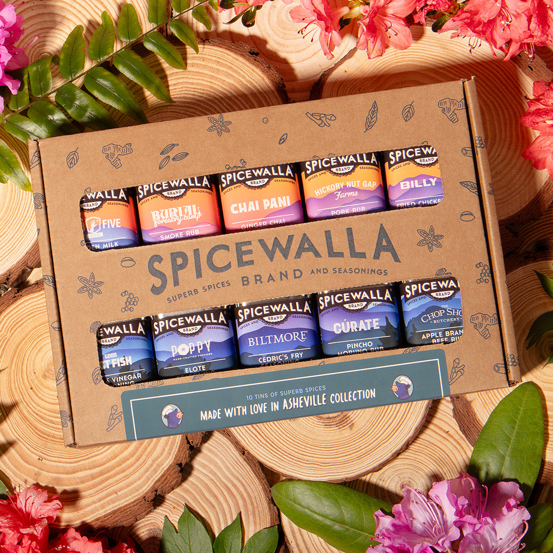 Spicewalla spice jars in a cardboard box surrounded by pink flowers and green leaves on wooden planks.