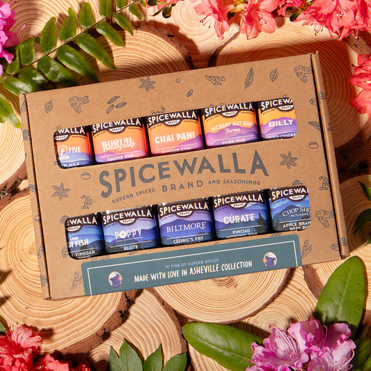 Spicewalla spice jars in a cardboard box surrounded by pink flowers and green leaves on wooden planks.