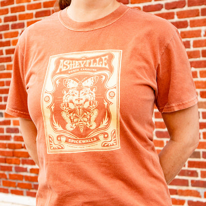 Person wearing a terracotta-colored t-shirt with a graphic design in front of a brick wall.
