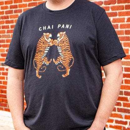 Person wearing a black t-shirt with tiger design and 'Chai Pani' text in front of a brick wall.