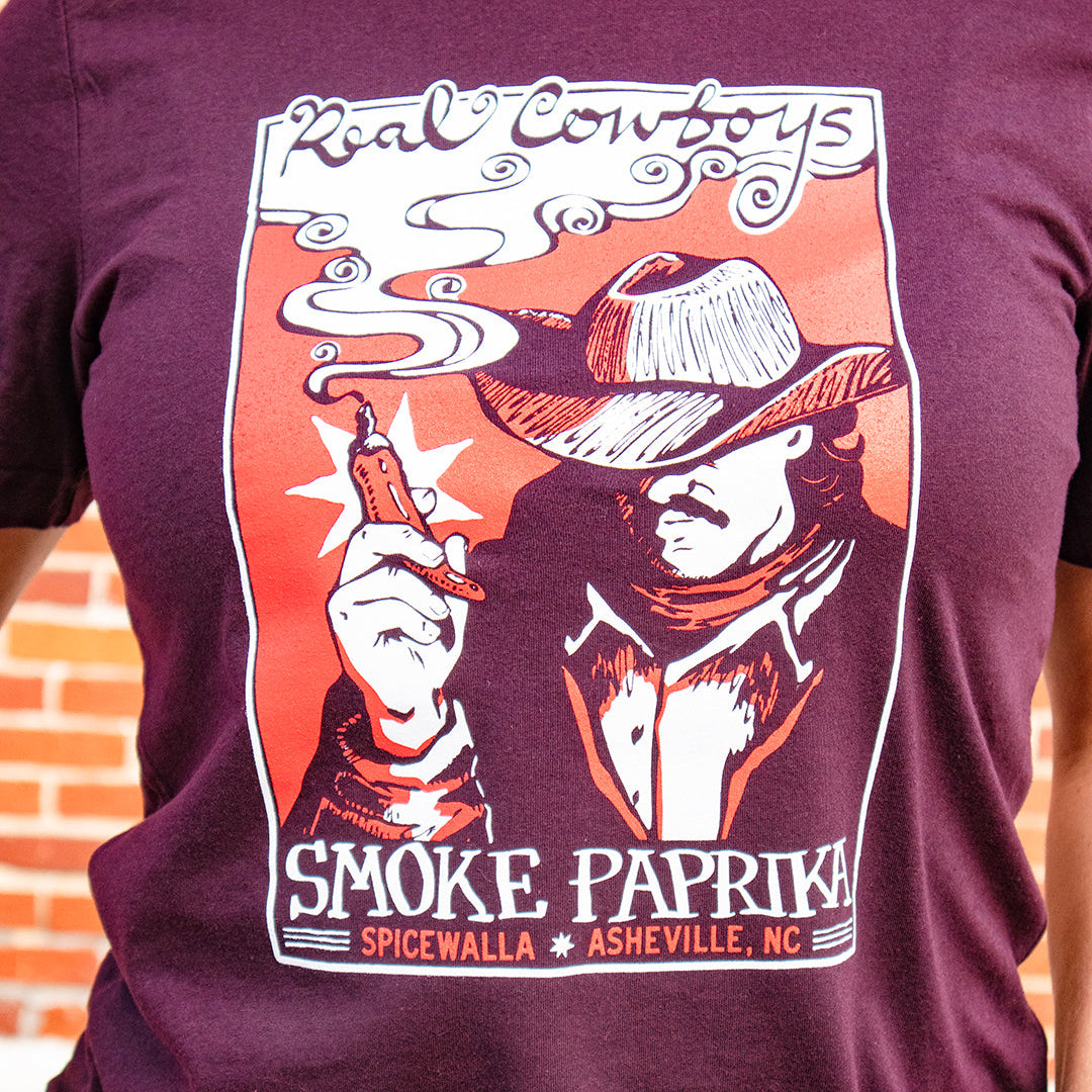 Maroon t-shirt with a graphic design of a cowboy smoking a cigarette, text 'Real Cowboys Smoke Paprika' on a brick wall background.