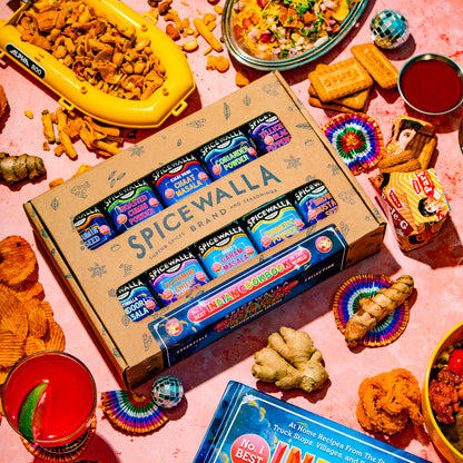 A styled flat-lay featuring a Spicewalla Indian Blend spice box, a copy of Meriwether's Best Indian Cookbook, and various colorful Indian dishes and ingredients on a pink surface.