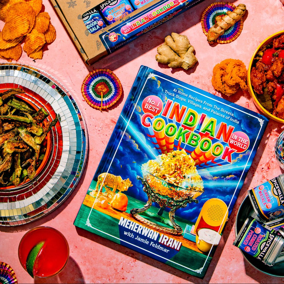 A styled flat-lay featuring a Spicewalla Indian Spicy Snack Box box and a Mehkiwin Iran 'Indian Cookbook' on a pink background surrounded by food items like chips, ginger, and colorful plates.