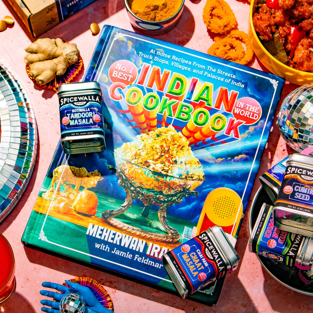 A colorful 'Indian Cookbook' book lies on a pink surface surrounded by food dishes, branded 'Spice Mall' tins, a disco ball, and other party props in a bright styled flat-lay scene.