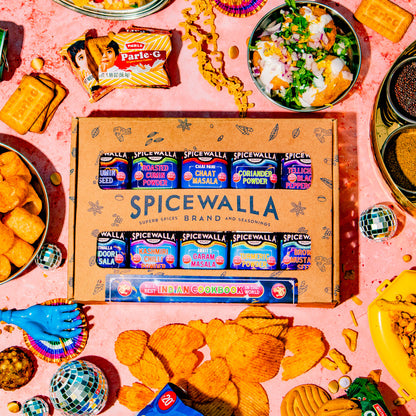 A Spicewalla brand snack box arranged on a pink surface surrounded by colorful snack props, including chips, dips, crackers, and small dishes. The central product box reads SPICEWALLA and shows multiple spice-flavored snack packs inside.