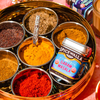A close-up styled product shot of a brass spice tray filled with assorted whole and ground spices, with a Spicewalla Garam Masala tin placed on top, against a pink background with decorative festive elements.
