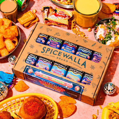 A Spicewalla gift box of chocolate-covered biscuits is displayed in a vibrant, festive flat-lay arrangement surrounded by Indian snacks, chutneys, and party decorations on a pink background.