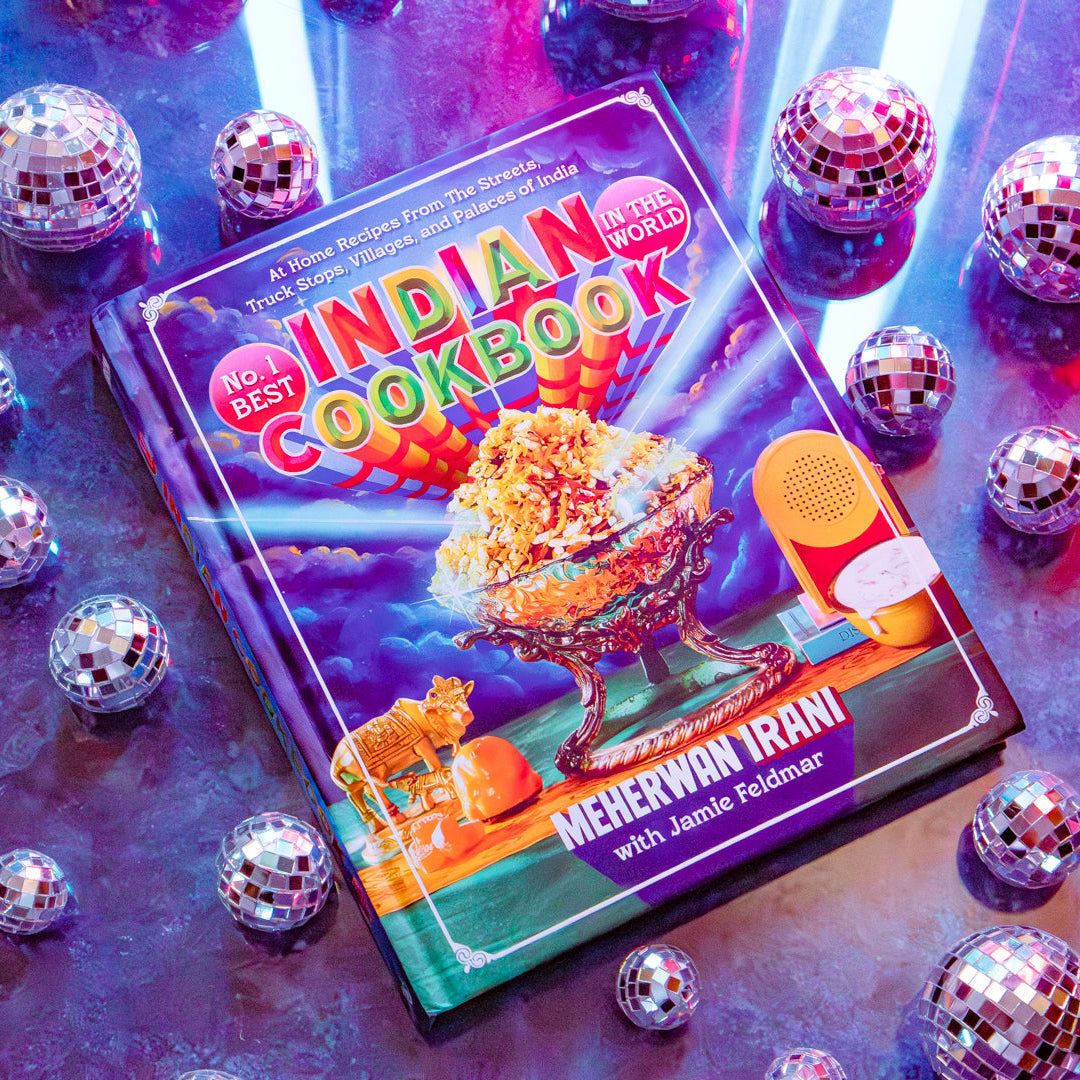A colorful hardcover book titled "No. 1 Best Indian Cookbook" lies on a reflective surface surrounded by many silver disco balls under neon pink, blue, and purple lighting.