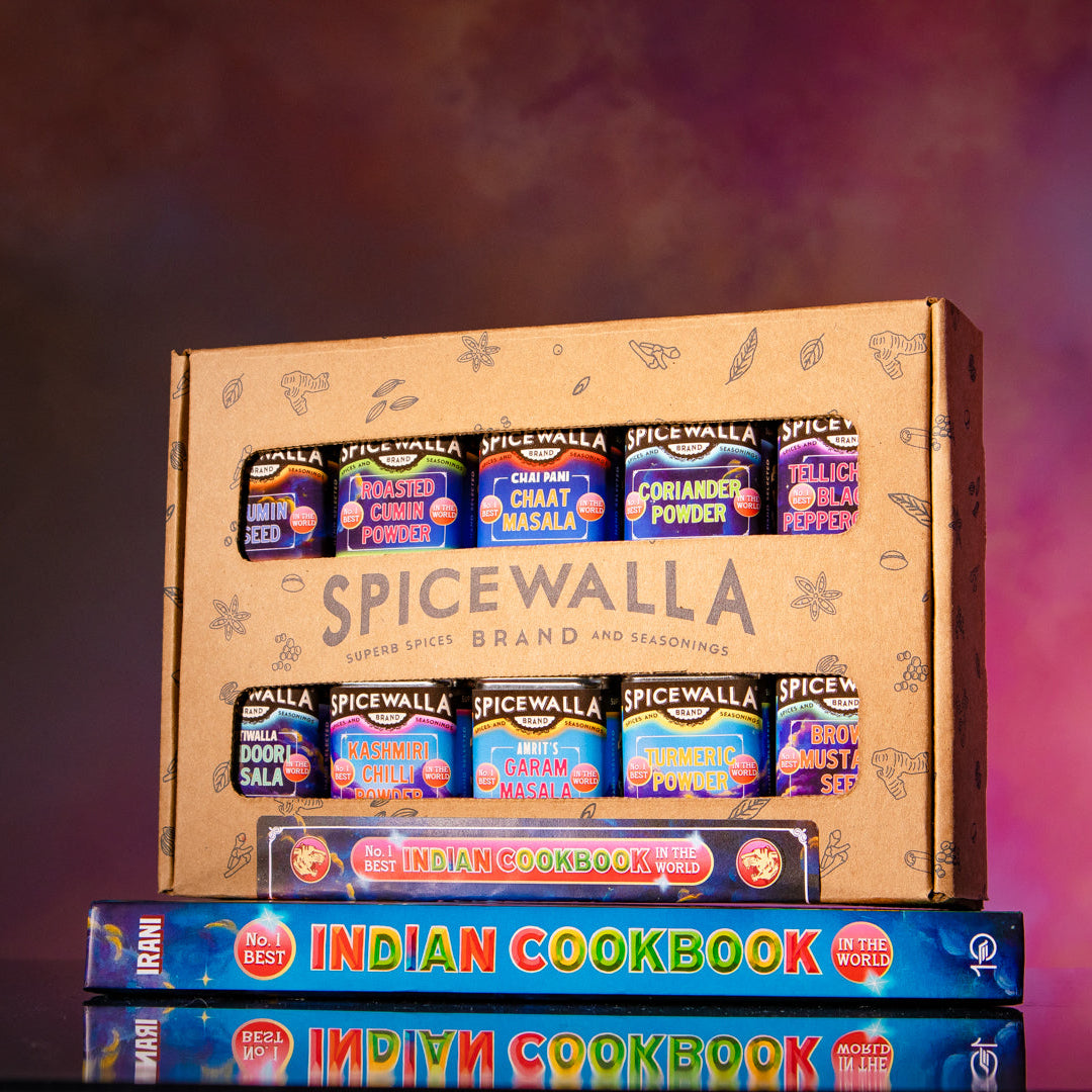 A SpiceWalla gift box filled with assorted spice jars, displayed on top of Indian Cookbook volumes against a smoky purple and pink studio background.