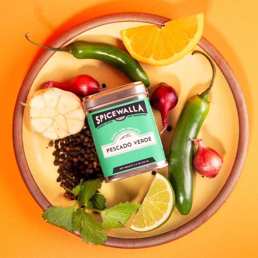Spicewalla Pescado Verde spice can surrounded by peppers, garlic, lemon, and lime on a yellow plate with an orange background.