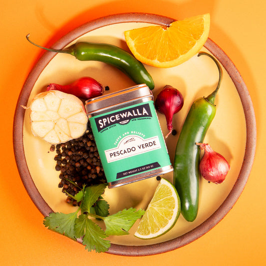 Spicewalla Pescado Verde spice can surrounded by peppers, garlic, lemon, and lime on a yellow plate with an orange background.