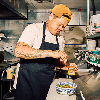 Person in a kitchen wearing an apron and yellow cap, preparing food.