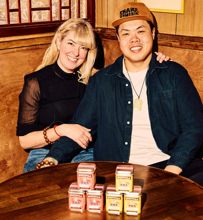 Two people sitting at a wooden table with small boxes in front of them, in a room with wooden paneling and a framed picture on the wall.