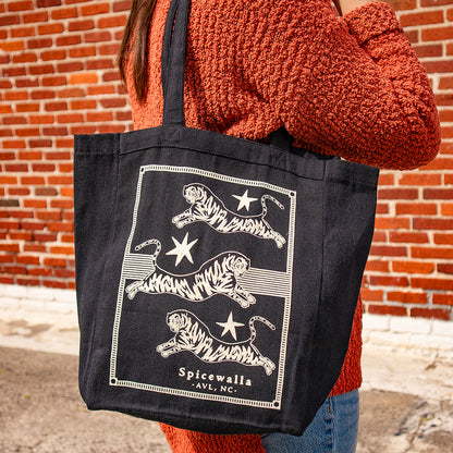 Person holding a black tote bag with tiger design and 'Spicewalla' branding against a brick wall.