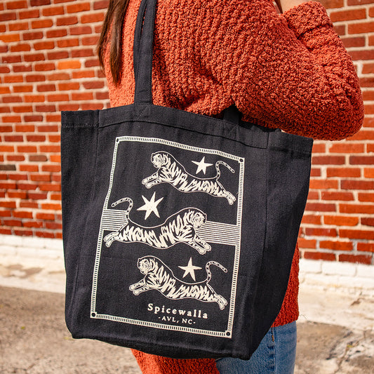Person holding a black tote bag with tiger design and 'Spicewalla' branding against a brick wall.